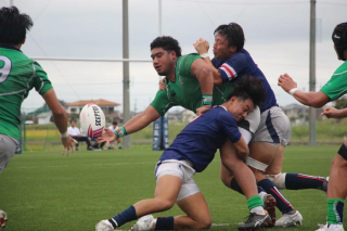 ラグビーの試合中、緑色のジャージを着た選手がボールを保持し、青いジャージの相手選手たちと激しくぶつかり合っている瞬間