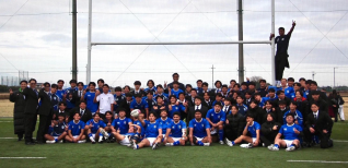 ラグビーのゴールポストがあるグラウンドで、青いユニフォームを着た選手たちとスーツ姿のスタッフが集合写真を撮っています