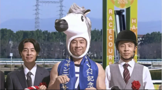競馬場を背景に3人の男性が並ぶ。中央の男性は馬の着ぐるみを着て青いタスキをかけ、両脇の男性はスーツとジョッキーの格好をしている。何かのイベントや宣伝の一コマのようだ。